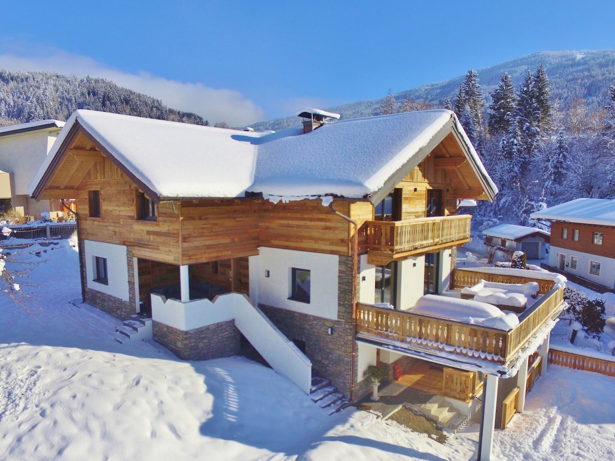 A charming holiday home is set against a winter landscape, featuring a wooden exterior with a snow-covered roof. The structure showcases multiple balconies and a grand staircase leading to the entrance. Surrounding the home is a blanket of fresh snow, with trees in the background.