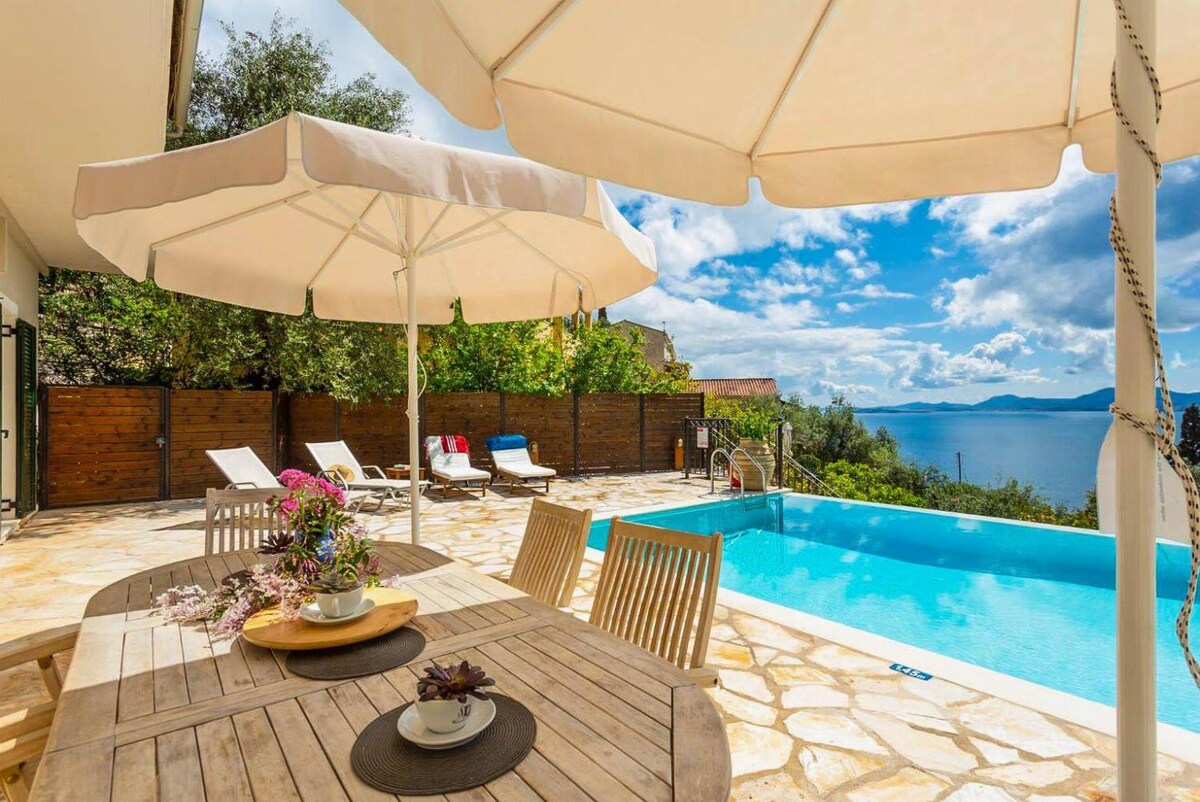 A sun-drenched terrace features a dining table surrounded by umbrellas, with a view of the private pool and the sea in the distance. Comfortable sun loungers are positioned nearby, and the area is adorned with colorful flowers, enhancing the outdoor space.