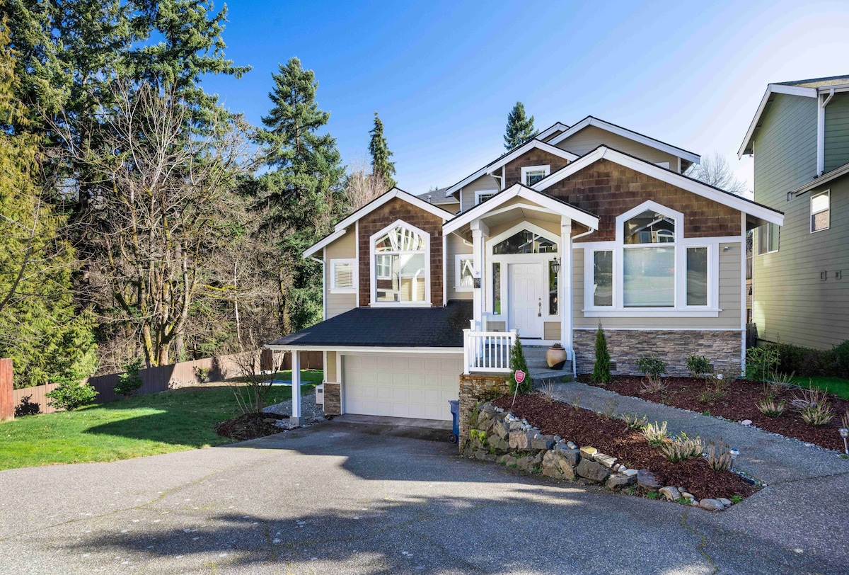 The exterior of the home is showcased, featuring a multi-level structure with warm wood tones. A welcoming front porch is visible, surrounded by landscaped gardens and lawn. Trees are observed in the background, enhancing the natural setting.