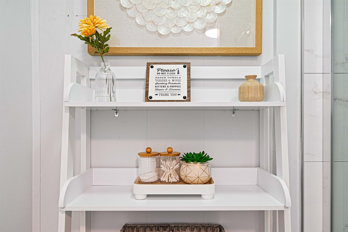 A white shelving unit is positioned against a light-colored wall, featuring decorative items. A small flower vase holds an artificial yellow flower, while a framed sign is displayed above. A wooden container, two glass jars, and a green plant with a textured pot are arranged neatly on the bottom shelf.