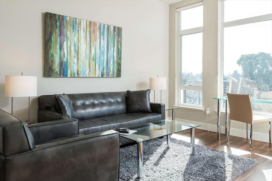 A modern living area features a sleek leather sofa and armchair, paired with a glass coffee table. Natural light floods the space through oversized windows, enhancing the colorful artwork displayed on the wall. A dining chair and table are positioned nearby, offering additional seating.