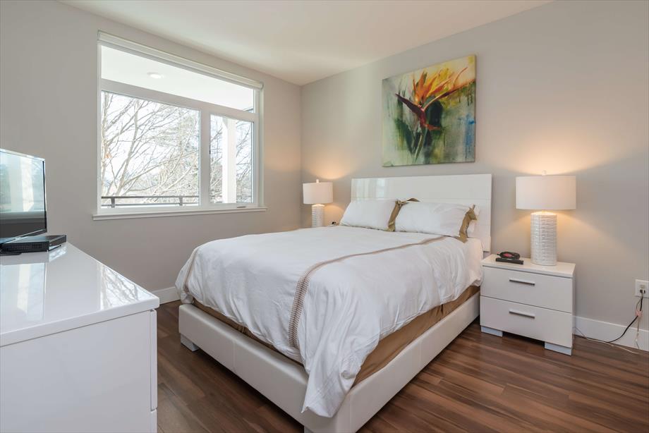 A bright bedroom features a spacious bed with a neutral-toned blanket, complemented by two bedside lamps. A modern piece of artwork adorns the wall above the bed. Ample natural light enters through oversized windows, and hardwood flooring enhances the room's contemporary appeal.