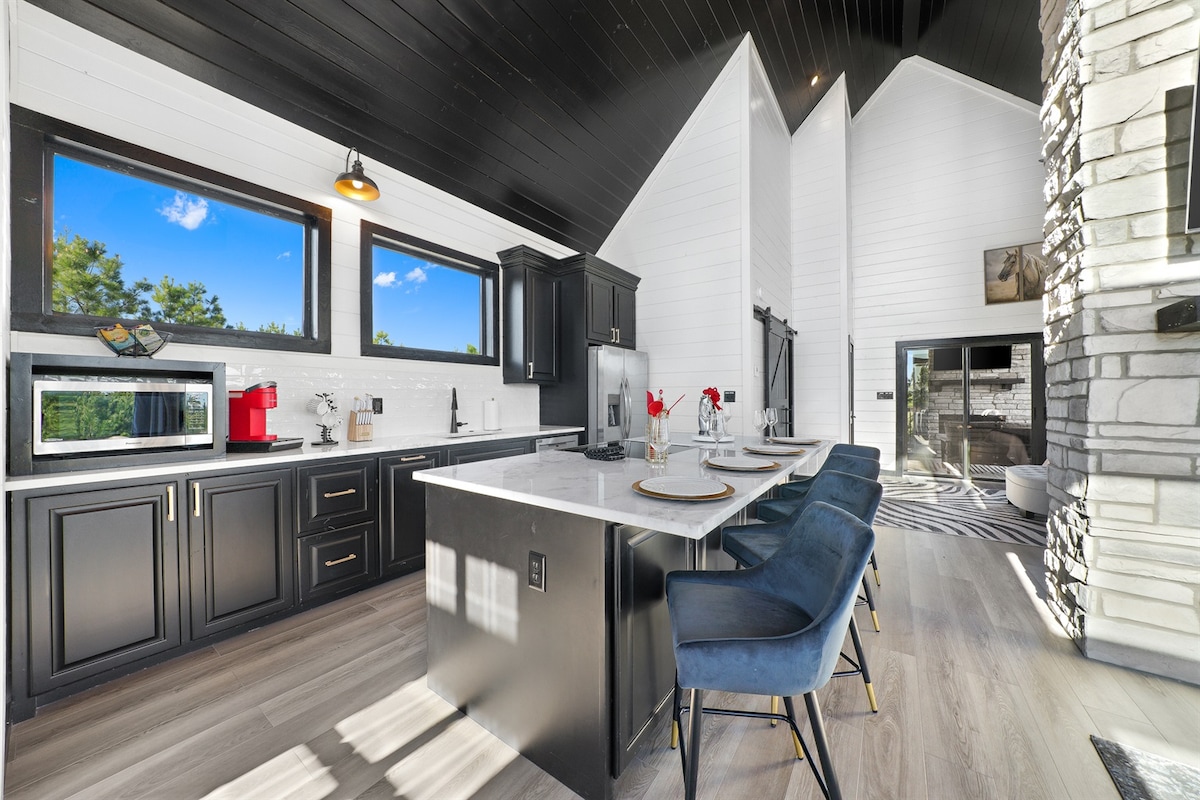A modern kitchen space features dark cabinetry and a white countertop, with three stylish blue barstools positioned at the island. Large windows provide natural light and showcase the surrounding view. A sleek stove and a microwave are integrated into the cabinetry, enhancing functionality.