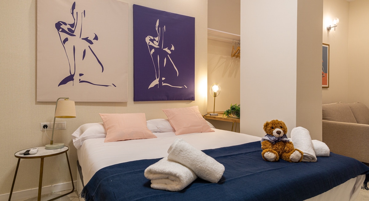 A spacious bed is positioned against the wall, accompanied by two framed artworks. Soft pink pillows adorn the bed, while neatly rolled towels are arranged at the foot. A decorative teddy bear sits nearby, providing a comforting touch to the inviting atmosphere.