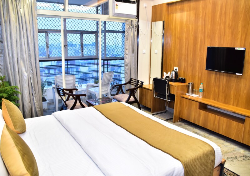 A spacious room features a large bed with neutral bedding, complemented by golden accent pillows. A wooden wall unit holds a flat-screen TV, and a desk is positioned nearby. A seating area with two chairs and a table is visible through floor-to-ceiling windows, offering city views.