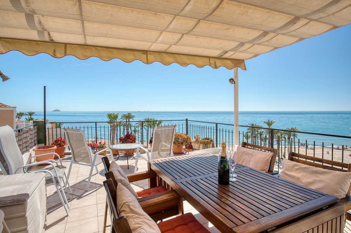 A spacious terrace features a wooden dining table surrounded by several chairs, complemented by comfortable seating areas. Potted plants add a touch of greenery, while the Mediterranean Sea is visible in the background. An awning provides shade, offering an inviting outdoor space.