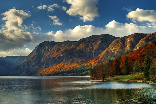 A serene lake is bordered by colorful autumn foliage on the surrounding mountains. The water reflects the sky and the landscape, while puffy clouds drift overhead. The scenery captures a tranquil and natural setting, inviting relaxation and exploration.