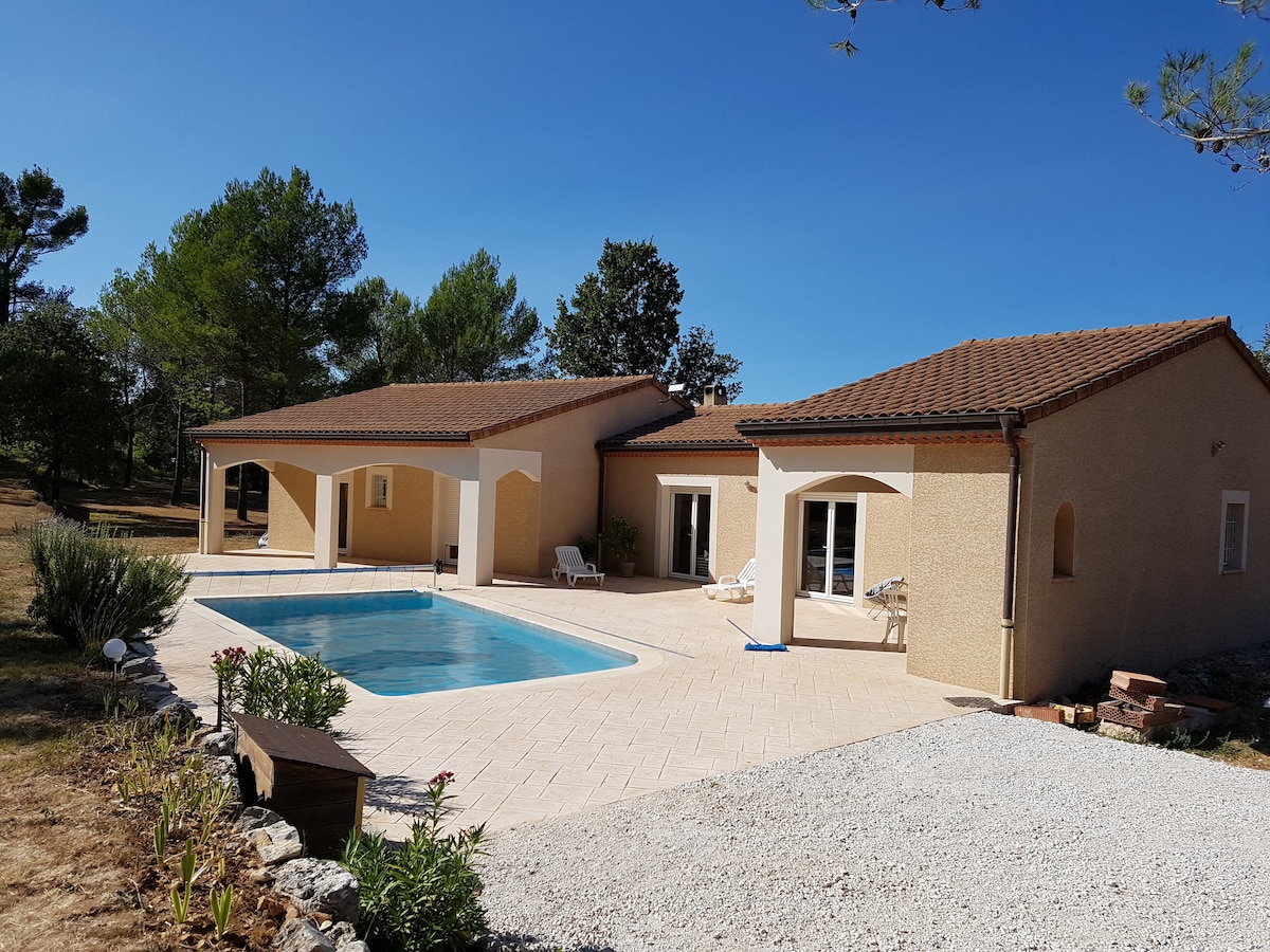 A sunlit exterior showcases a spacious house with a tiled patio surrounding a swimming pool. Lush greenery from nearby trees complements the serene outdoor setting, while a well-maintained garden enhances the tranquil atmosphere. The structure features inviting entryways and large windows for natural light.