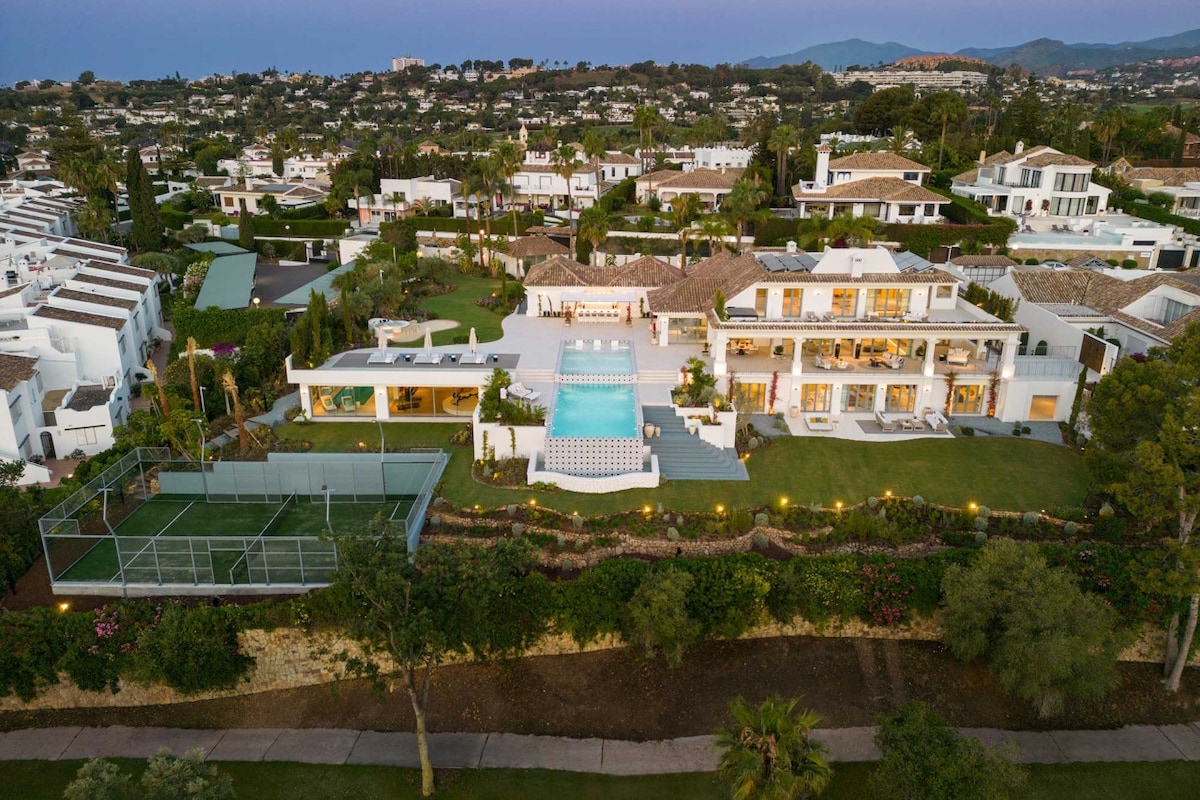 An aerial view of the luxurious villa reveals a spacious outdoor area, featuring well-maintained gardens and a striking pool area with a patterned tiled pool. A private padel court is also visible, highlighting the expansive layout and welcoming atmosphere of the property.