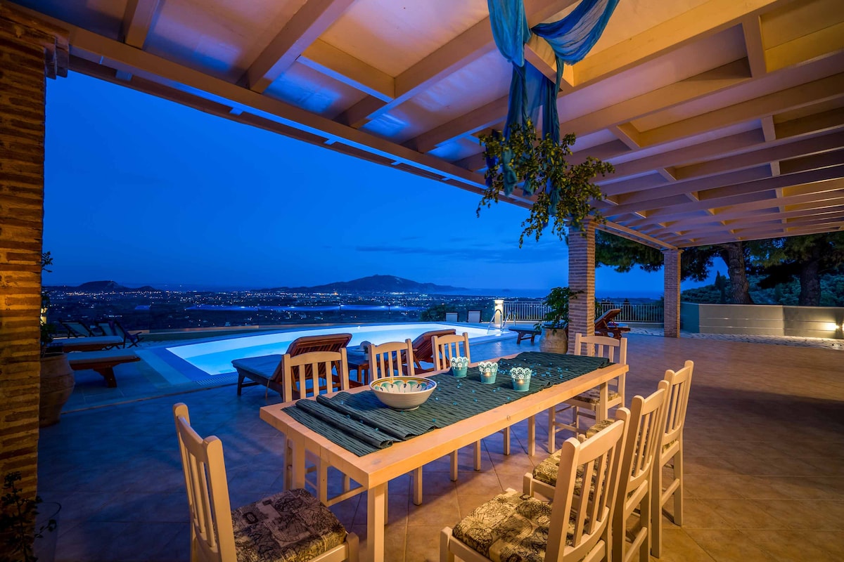 An outdoor dining area is presented with a large table and several chairs, set against a backdrop of vibrant blue dusk. The space is adorned with greenery and features views of the illuminated swimming pool and distant landscape, enhancing the evening ambiance.