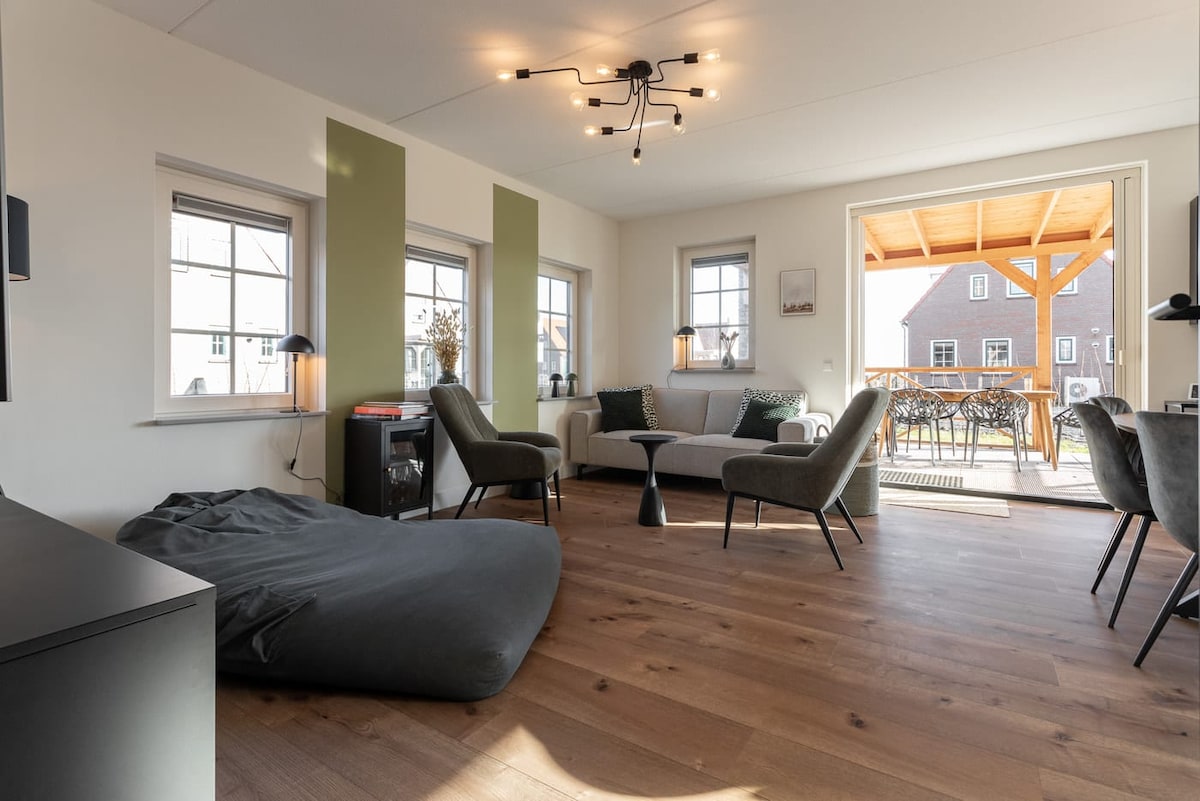 A bright living area features a mix of seating options, including a cozy bean bag and stylish armchairs. Natural light streams through large windows, and doors open to an outdoor seating area. A modern light fixture hangs from the ceiling, adding a contemporary touch.