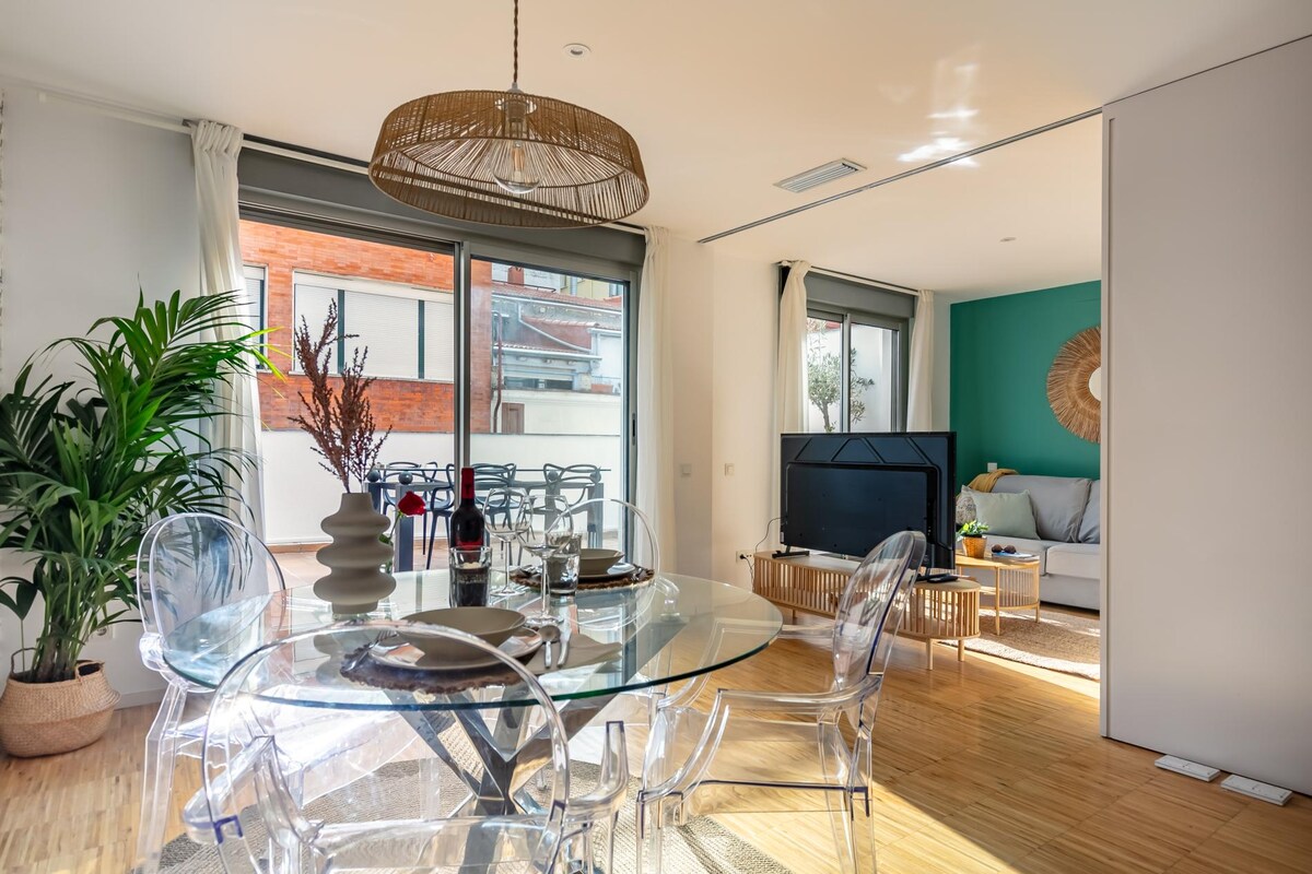 An open-plan living space features a circular glass dining table surrounded by transparent chairs. A modern TV unit is visible, with a cozy seating area in the background. Large windows allow natural light to fill the room, enhancing the connection to the outdoor terrace.