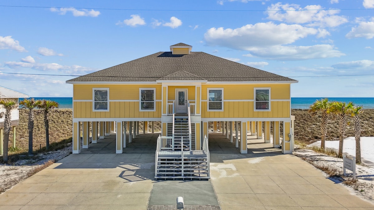 A spacious beach house with a bright yellow exterior is set against a clear blue sky, showcasing a large elevated porch. Palm trees line the property, while the Gulf of Mexico can be seen in the background, emphasizing the coastal location.