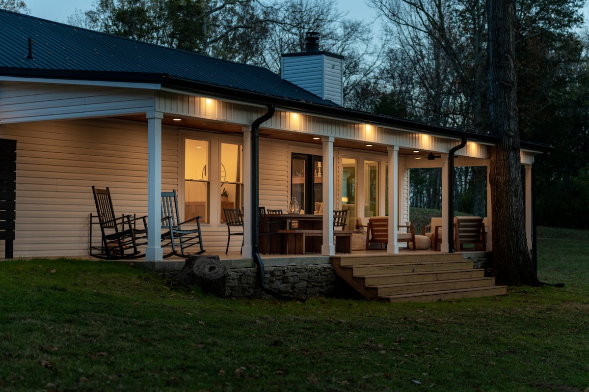 An evening view of the home’s exterior showcases a covered porch with rocking chairs and wooden seating. Warm light reflects from the windows, creating a welcoming ambience against the backdrop of trees and a grassy yard.
