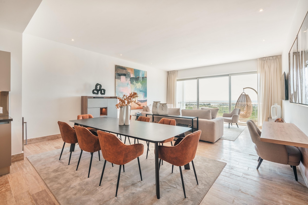 A spacious living area is highlighted by a long dining table surrounded by six comfortable chairs. A large abstract artwork adorns one wall, while natural light filters through the floor-to-ceiling windows, providing views of the surrounding landscape. A cozy seating area is situated to the right.