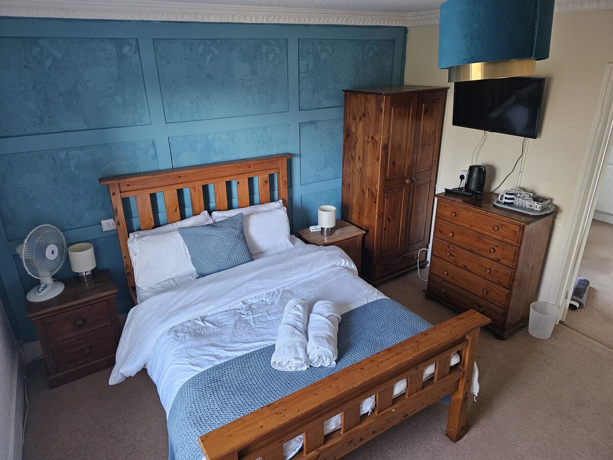 A cozy bedroom is furnished with a wooden bed, paired with a light blue blanket and fresh towels. Wood dressers and bedside tables offer storage, while a television is mounted on the wall. A standing fan enhances comfort in the well-lit space.