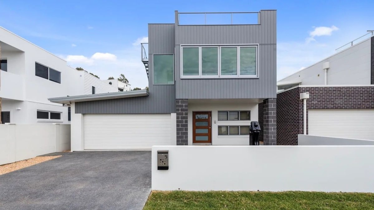 The modern facade of the residence is showcased with clean lines and large windows. A two-car garage is visible, complemented by a path leading to the entrance. A smooth driveway and a patch of grass add a welcoming touch to the exterior.