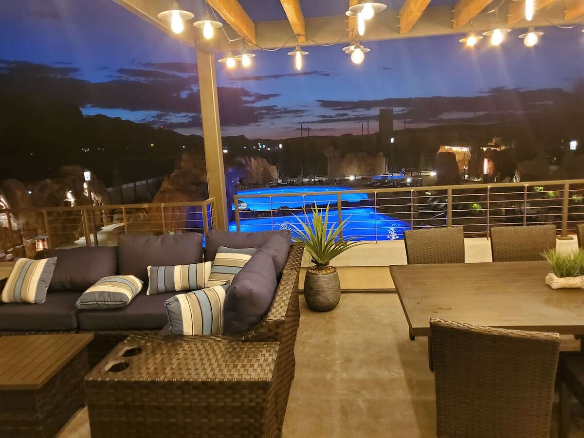 A covered terrace features comfortable seating with blue and white accent pillows, along with a dining area. The view includes resort-style pools illuminated in blue under evening skies, while warm string lights create a cozy ambiance.