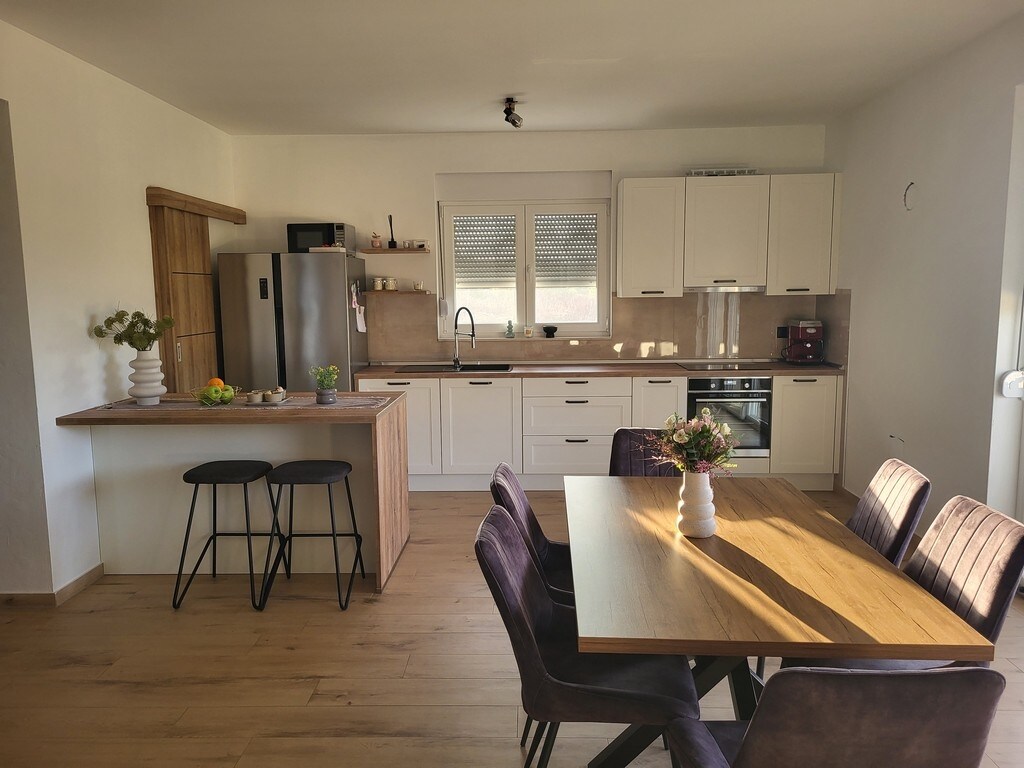 A modern kitchen is presented, featuring white cabinetry and a spacious countertop. An island with two stools is visible, along with a dining table surrounded by four upholstered chairs. Natural light enters through large windows, enhancing the warm wood flooring.