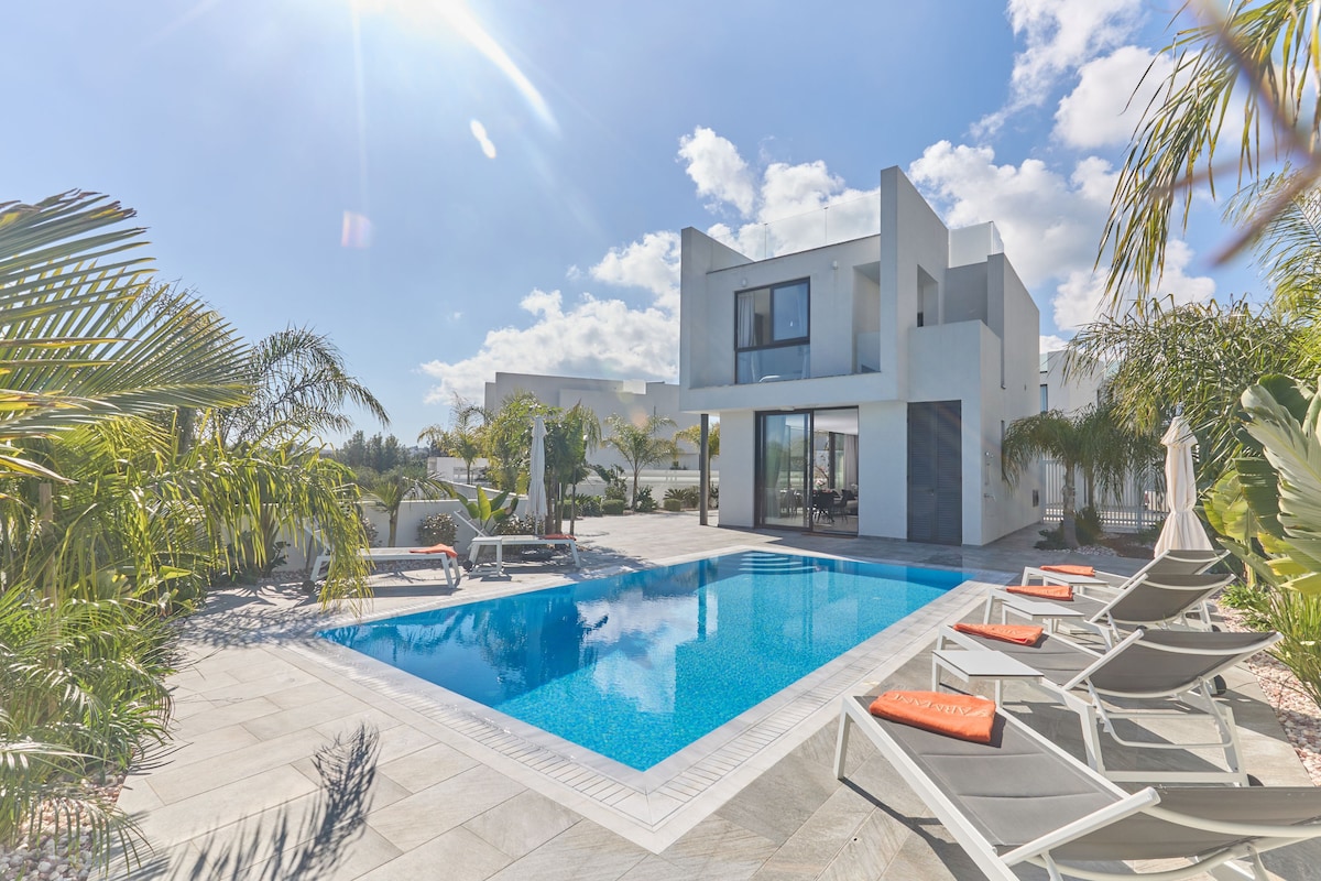 A modern villa with a sleek design is surrounded by palm trees, featuring a private pool with clear blue water. Comfortable sunloungers are positioned nearby, enhancing the outdoor relaxation area. Large windows reflect the sunny sky, creating an inviting atmosphere for leisure.