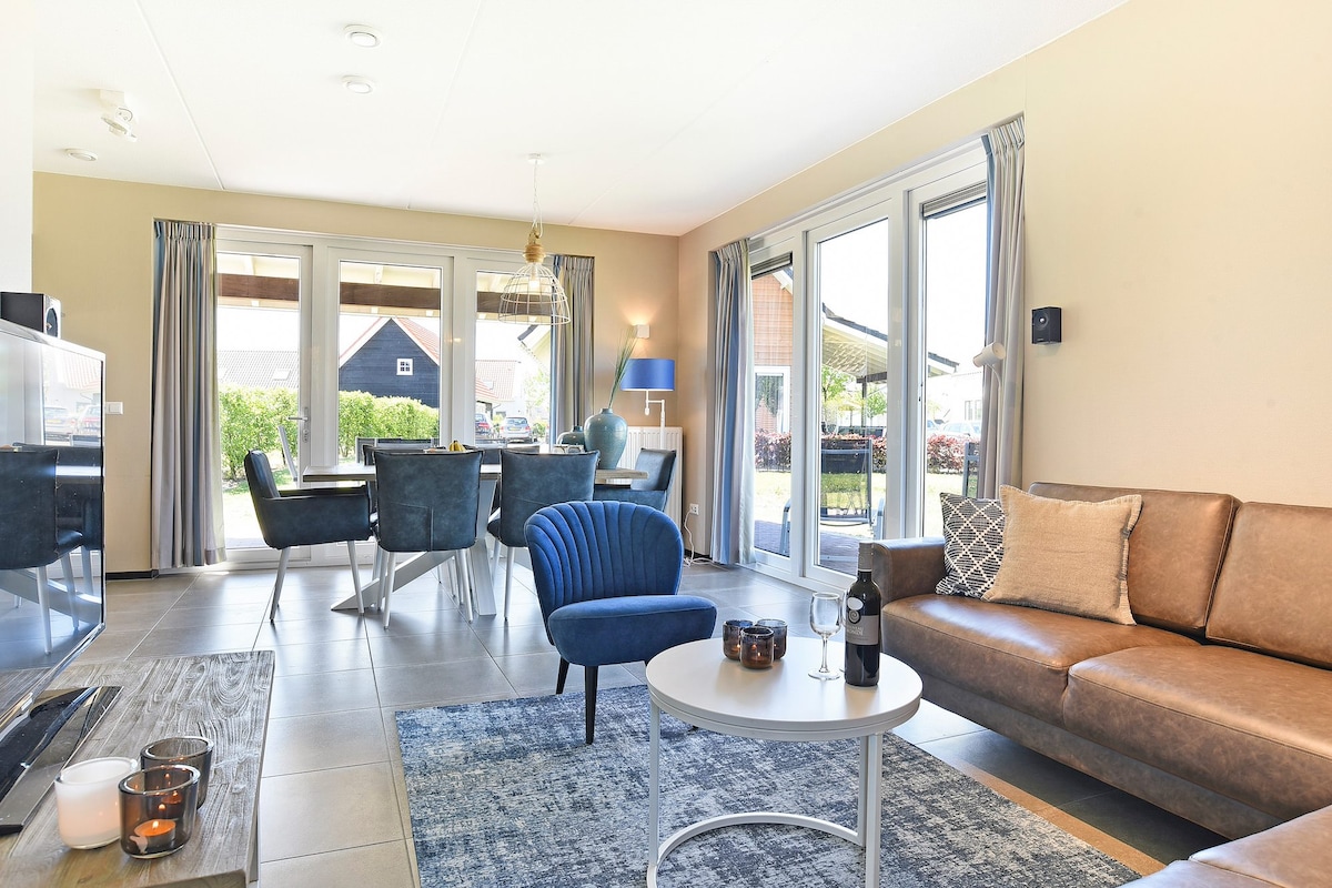 An open-plan living area is featured, showcasing a comfortable brown sofa and a blue accent chair. A circular table is set for dining, surrounded by chairs. Large glass doors provide a view of the outdoor space, allowing natural light to fill the room.