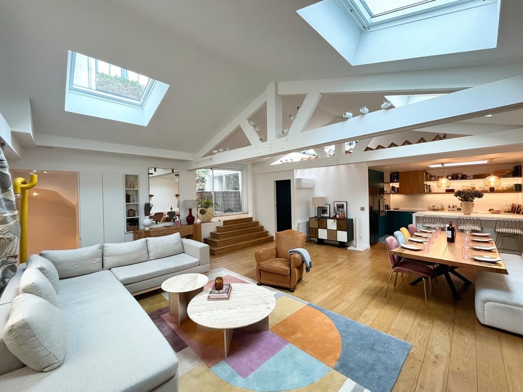 A spacious living area features high ceilings with skylights, allowing natural light to fill the room. A large sectional sofa and armchair are positioned around a circular coffee table atop a colorful area rug. A dining table for 8-10 people is set against a modern kitchen backdrop.