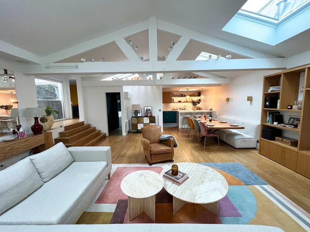 The spacious living area features a modern design with high ceilings and wood flooring. A large light-colored sofa faces two marble coffee tables, while a dining area with seating for eight is visible in the background. Natural light floods through a skylight, enhancing the open layout.