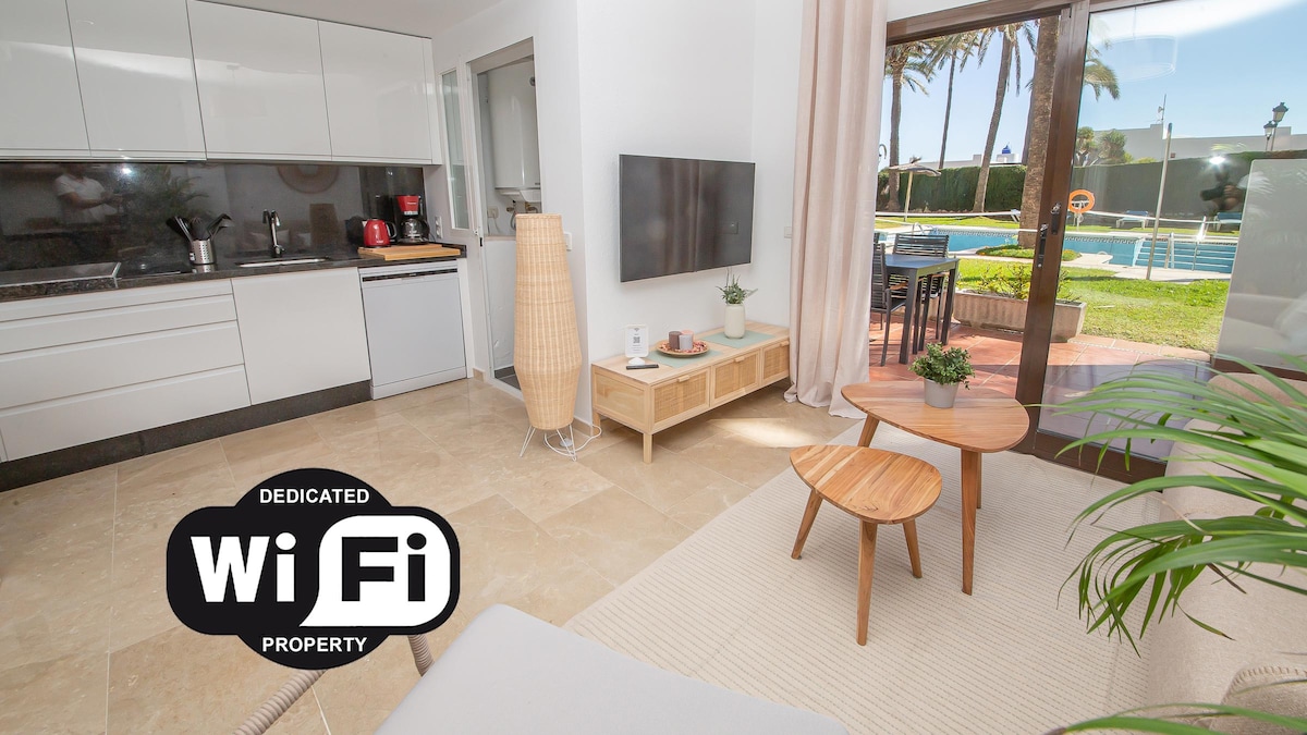 An open-plan kitchen and lounge area is visible, featuring modern cabinetry and appliances. A comfortable coffee table is positioned between two wooden chairs. Large glass doors provide direct access to the terrace and pool area, enhancing the sense of spaciousness.