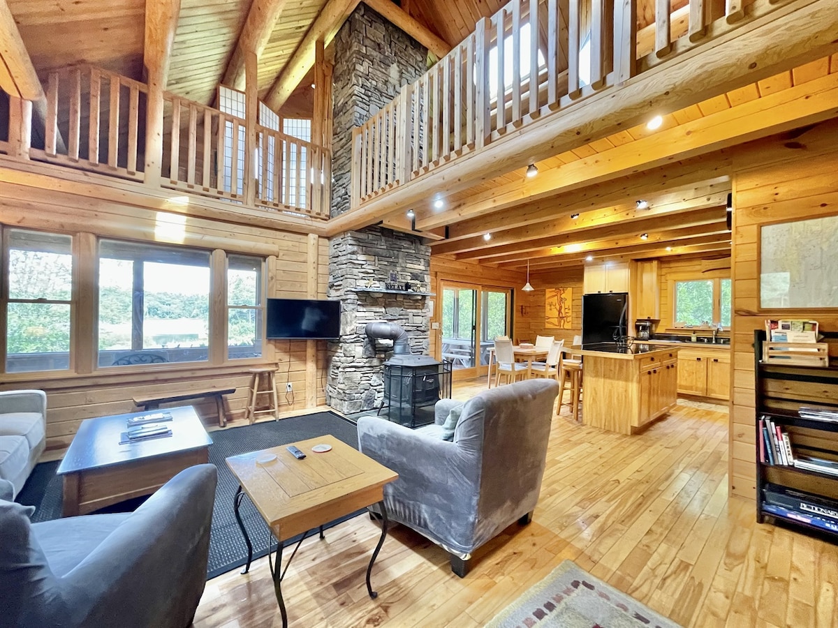 An open living space features a high wooden ceiling and stone accents. Large windows allow natural light and showcase views of the lake. Comfortable seating arrangements include a grey sofa and wooden coffee table, with a dining area visible in the background.