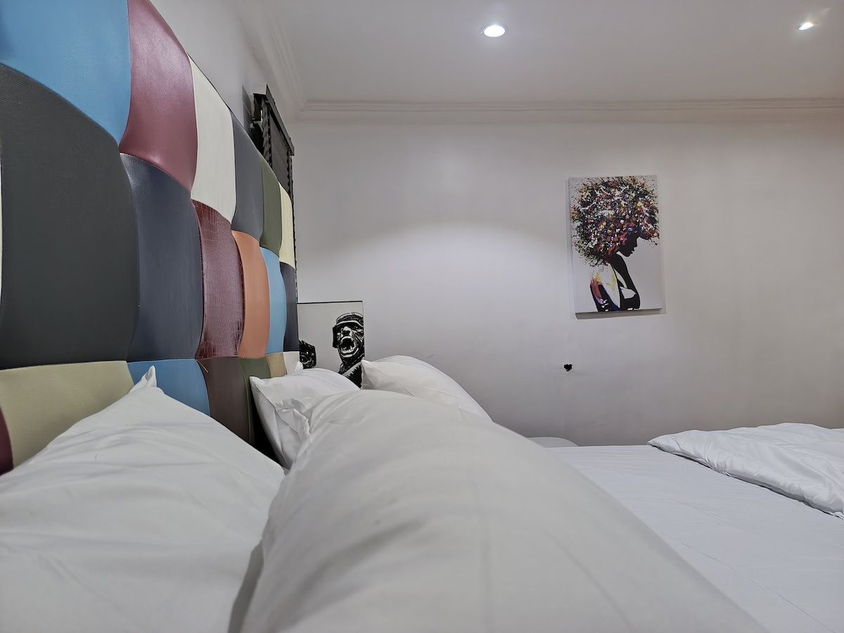 A vibrant headboard with multicolored patches is featured above the spacious beds, providing a modern touch. A striking artwork of a woman can be seen on the wall, while soft pillows are arranged on the beds, creating a restful ambiance.