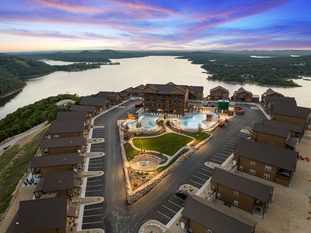 Aerial view showcases the lodge's layout amidst lush surroundings, highlighting the outdoor pool features and winding pathways. Table Rock Lake glistens in the background, with sunset hues painting the sky above the scenic complex.