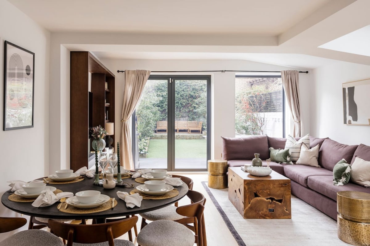 An inviting open-plan reception room features a circular dining table set with dinnerware, surrounded by upholstered chairs. A cozy sectional sofa is positioned along one wall, and large glass doors open to a green garden space, enhancing the natural light flowing into the room.