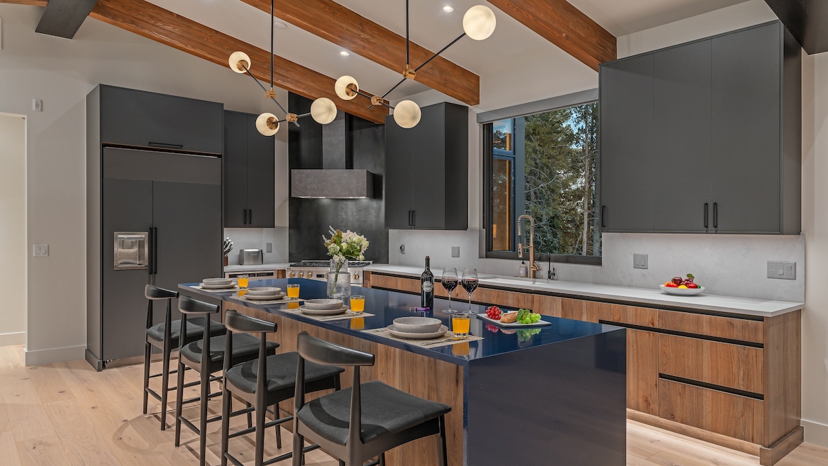 The kitchen features a modern design with sleek cabinetry and an expansive island. Four barstools are arranged around the island, complemented by flowers and colorful food items. Natural light flows in through the large window, illuminating the wooden beam accents overhead.