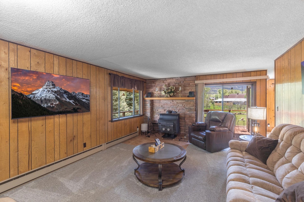 A cozy living room features wooden walls and a soft carpet. A large television is mounted above a brick fireplace. Comfortable seating includes a plush sofa and an armchair, creating a welcoming space. A window provides natural light and views of the surrounding scenery.
