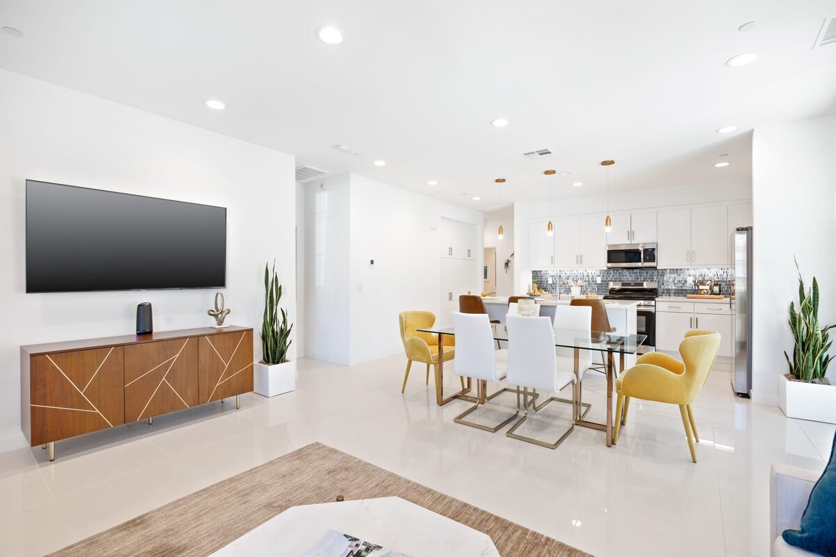 A modern living space showcases a mounted flat-screen TV above a stylish wooden console. A dining area features a glass table surrounded by six chairs, with two in bold yellow. The kitchen is visible in the background, accented by sleek finishes and pendant lighting.