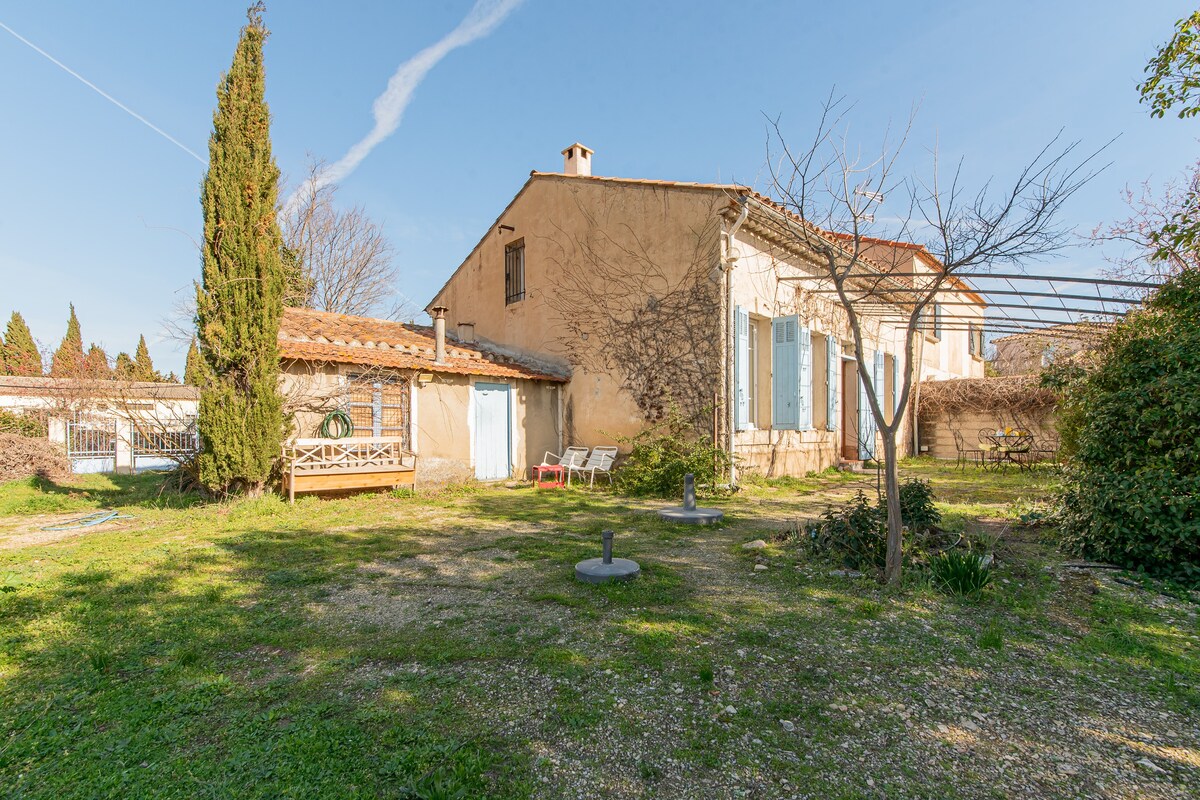 An expansive garden is showcased, featuring a mix of grass and gravel areas. A sunlit terrace is visible, accompanied by scattered seating options. Lush trees and shrubs surround the garden, creating a serene outdoor space. The house's rustic exterior with blue shutters is also part of the view.