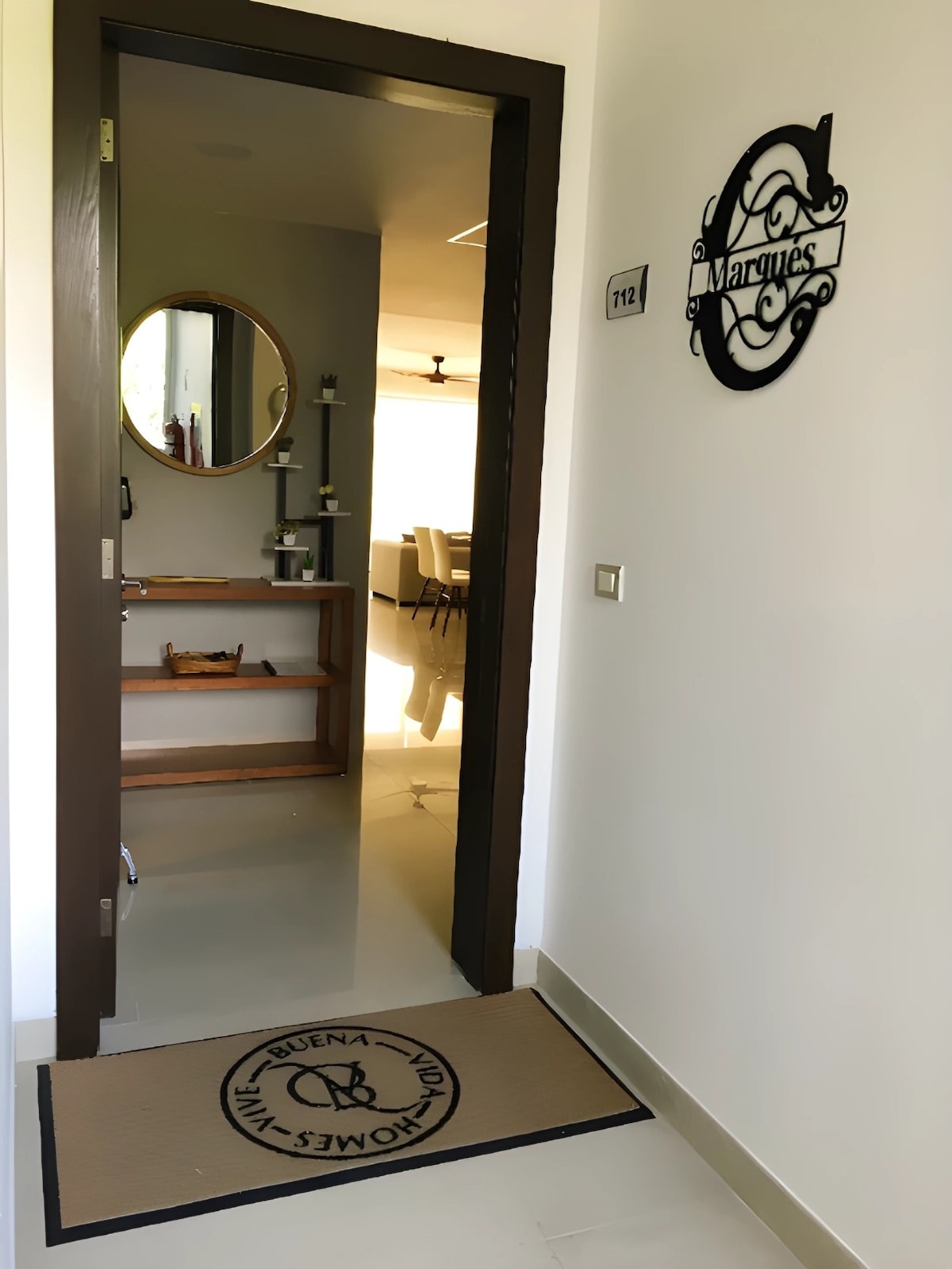 The image captures the entrance to Unit 112 of Casa Marquéz. A sleek door with an inviting sign is visible, along with a round mirror and a wooden shelf displaying decorative items. A welcome mat lies at the entrance, promoting warmth and hospitality.