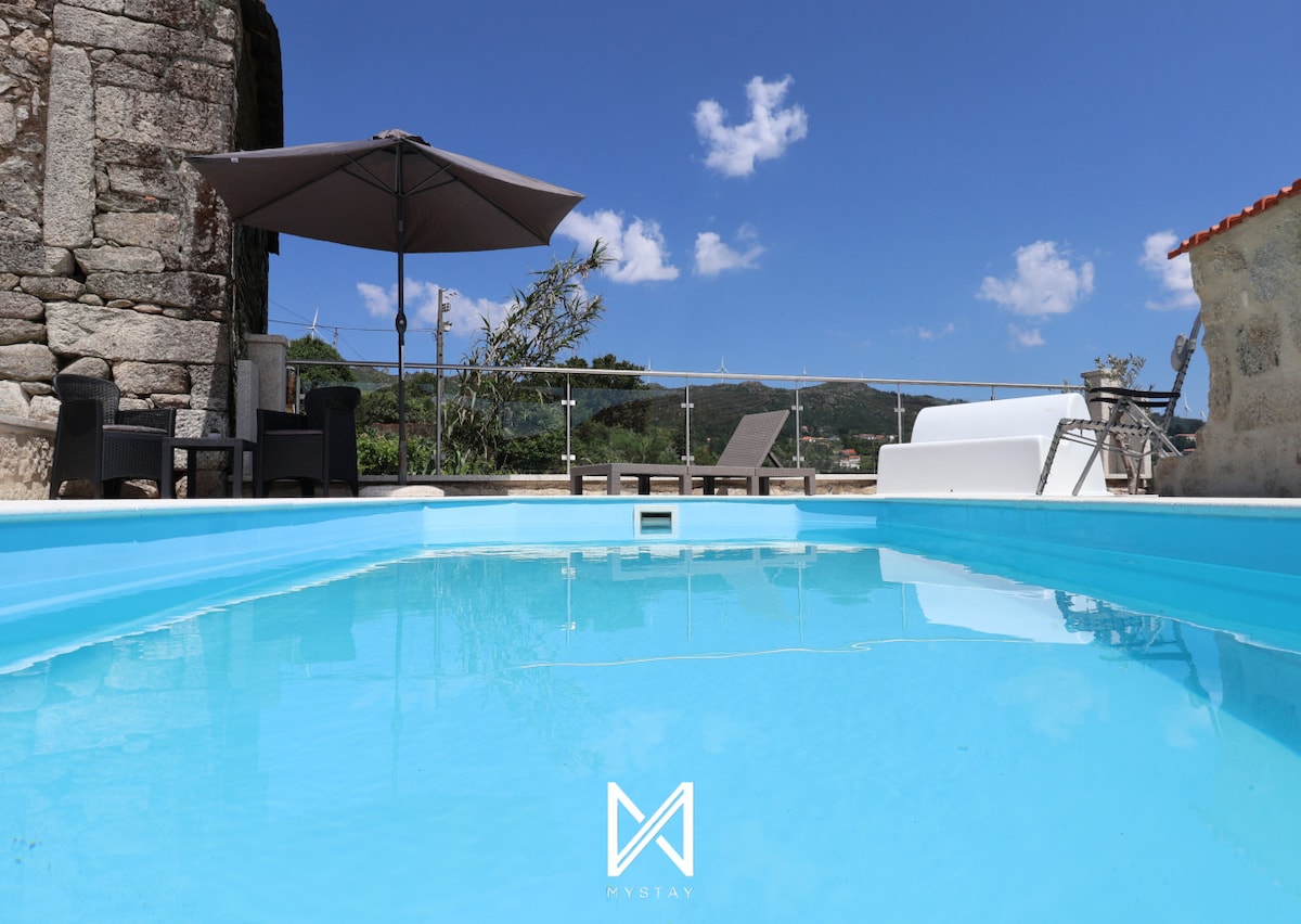 A private pool is visible, reflecting clear blue skies and surrounded by a sunlit deck. Lounge chairs are arranged nearby under a large umbrella, providing shaded seating. The backdrop features a mix of greenery and rustic stone architecture, enhancing the tranquil outdoor space.