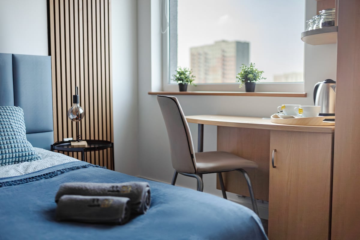 A modern guest room is presented, featuring a spacious bed with a textured blue blanket. A wooden desk and a chair are visible beside a window, adorned with small potted plants. A bedside table holds a lamp, and a tray with a kettle and refreshments is positioned nearby.