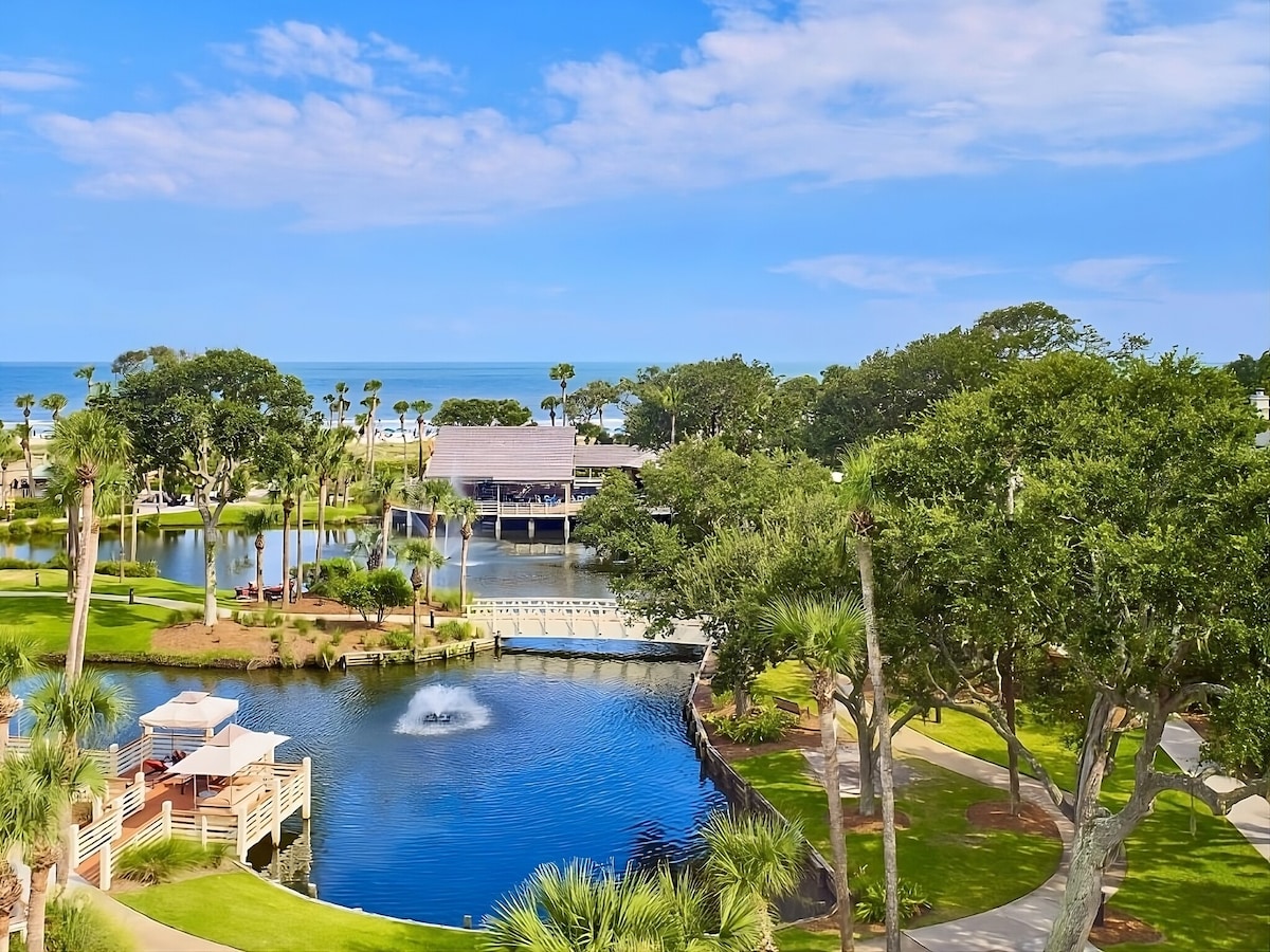 A scenic view captures the tranquil landscape featuring lush greenery and a serene pond with a gentle fountain. The backdrop reveals pristine beach access and the ocean beyond, complemented by the presence of palm trees and recreational facilities nearby.