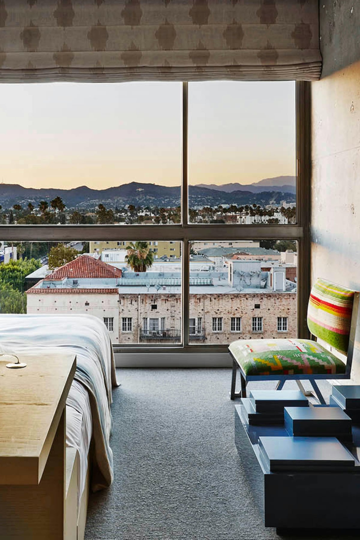 A spacious room features a King bed with neutral linens and a modern chair with a colorful pattern. Floor-to-ceiling windows provide views of the neighborhood and mountains, with soft shades partially drawn to filter light. A minimalist wooden table adds functionality.
