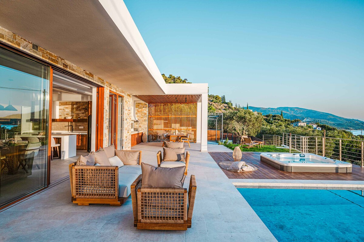 A spacious terrace is featured, showcasing comfortable lounge seating with neutral cushions arranged around a glass table. Nearby, a private hot tub sits beside an inviting infinity pool, with panoramic views of the surrounding landscape and coastline visible in the background.