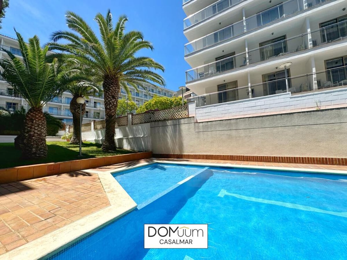 A clear swimming pool is bordered by palm trees, featuring a sunlit terrace with a tiled surface. The pool's refreshing blue water contrasts with the surrounding greenery and the clean lines of the residential building in the background.