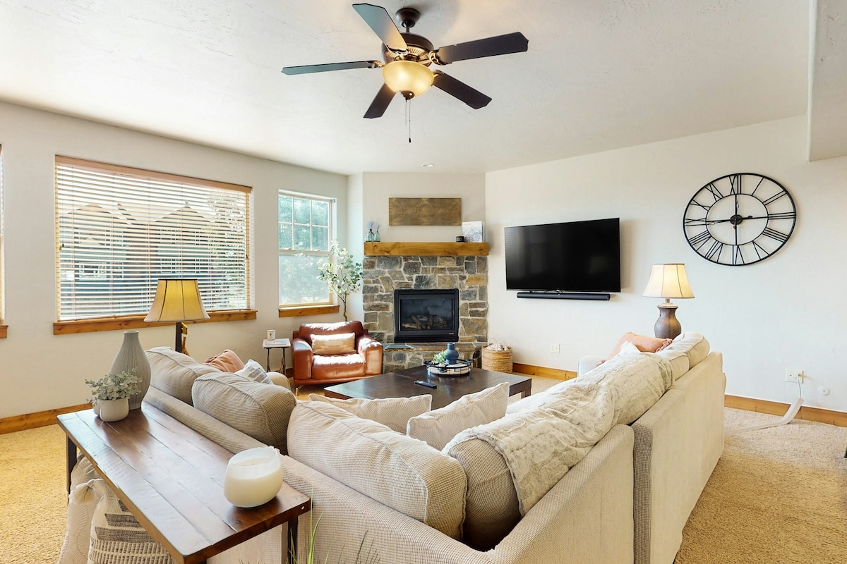 The living room is furnished with a large sectional sofa facing a stone fireplace. Natural light is provided by multiple windows, and a ceiling fan hovers above. A coffee table sits in the center, surrounded by a side table and a pair of armchairs.
