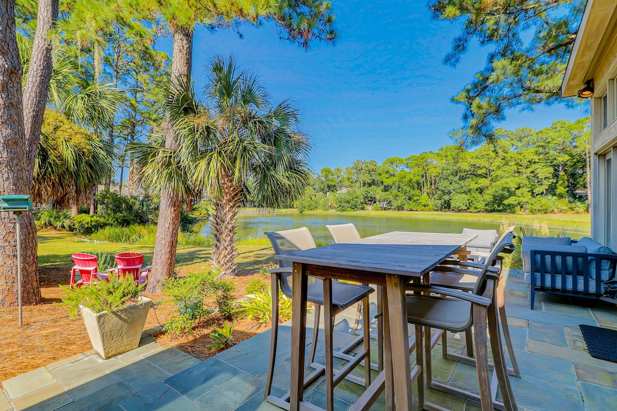 An outdoor dining area features a high table surrounded by bar stools, positioned on a stone patio overlooking a serene lagoon. Lush greenery and palm trees frame the view, while a casual seating arrangement sits nearby, inviting relaxation amidst nature.