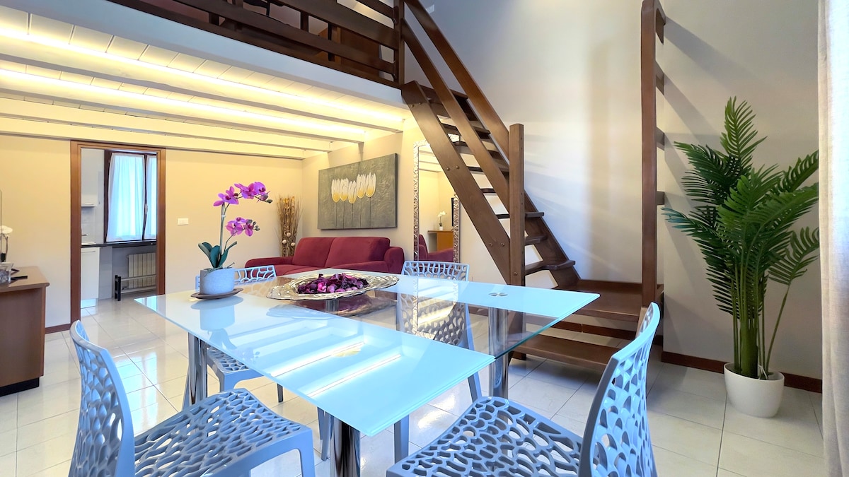 A spacious dining area is highlighted by a glass table surrounded by four white chairs. A vibrant orchid centerpiece enhances the setting. In the background, a bright living space features a maroon sofa, and a staircase leads to an upper loft area. Natural light filters through the windows.