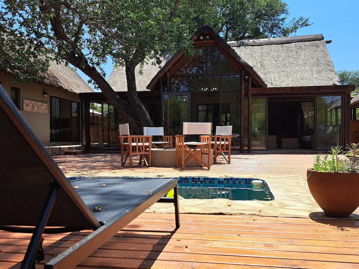 A sunlit outdoor deck features a small pool and a wooden table surrounded by four chairs. A spacious glass-fronted structure is visible in the background, framed by lush greenery and shaded by a thatched roof.