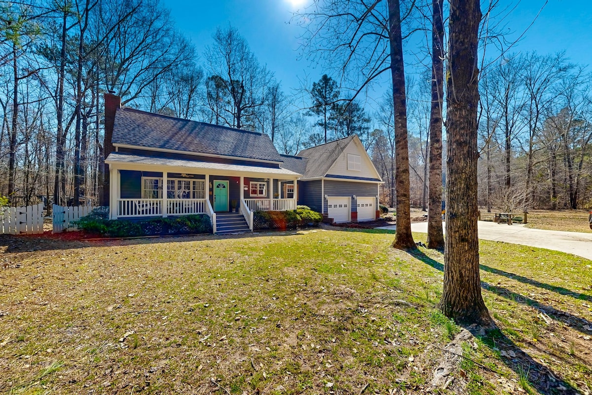 The exterior of a charming farmhouse-style home is depicted, featuring a front porch, a large driveway, and twin garages. The surrounding area is shaded by tall trees, with a green lawn showing traces of fallen leaves, creating a serene outdoor setting. Bright blue skies provide a sunny backdrop.