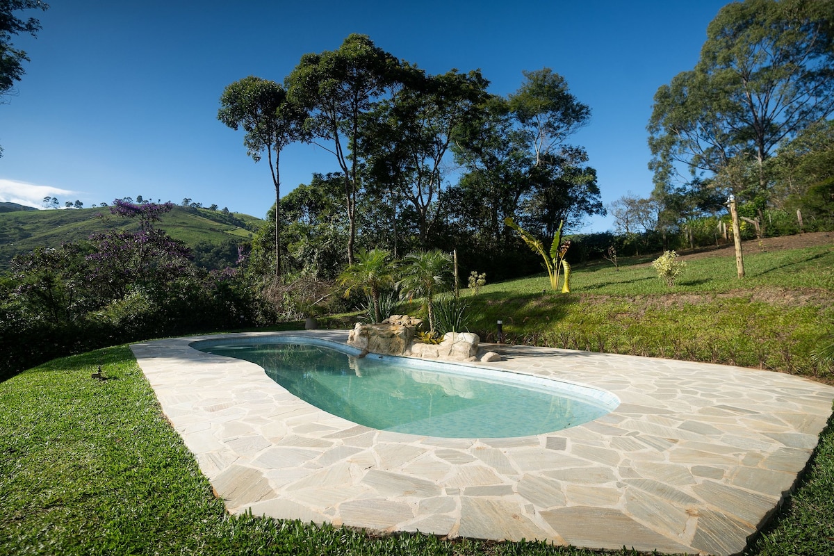 A natural-looking pool is surrounded by stone tiles, blending seamlessly with the landscaped garden. Lush greenery, including trees and plants, adds to the tranquil atmosphere. The serene setting is enhanced by distant hills under a clear blue sky.