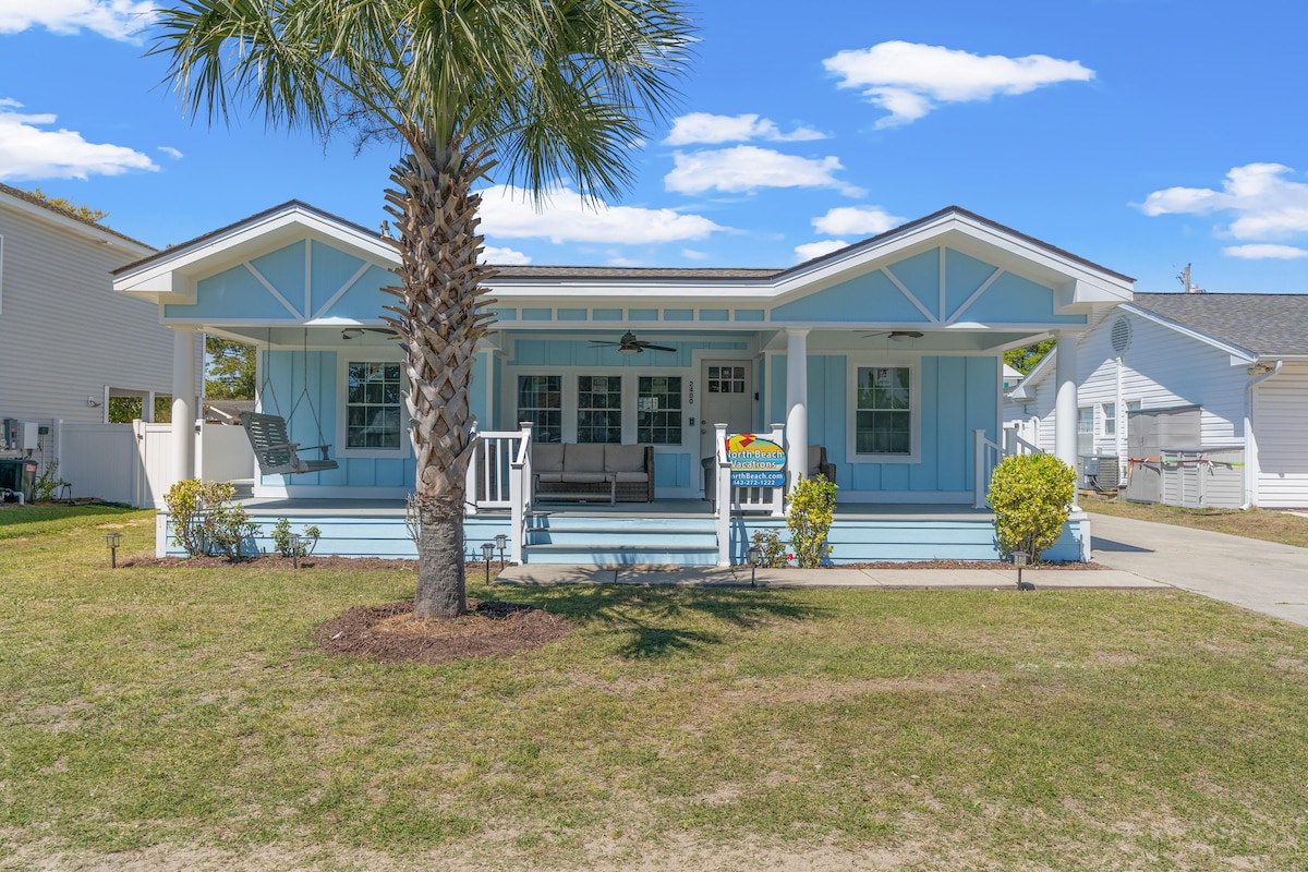 A charming beach house features a light blue exterior with a covered porch. A palm tree stands prominently in the front yard, surrounded by neatly trimmed grass and small shrubs. The porch includes seating arrangements, inviting relaxation in the coastal environment.